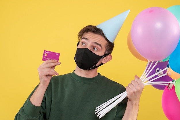 Front view young man with party cap and black mask holding card and balloons on yellow 