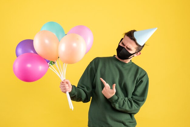 Front view young man with party cap and black mask pointing at balloons on yellow 