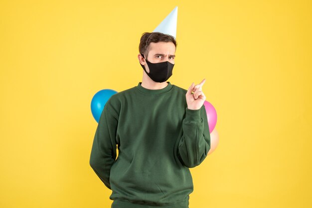 Front view young man with party cap and colorful balloons hiding balloons behind his back standing on yellow 