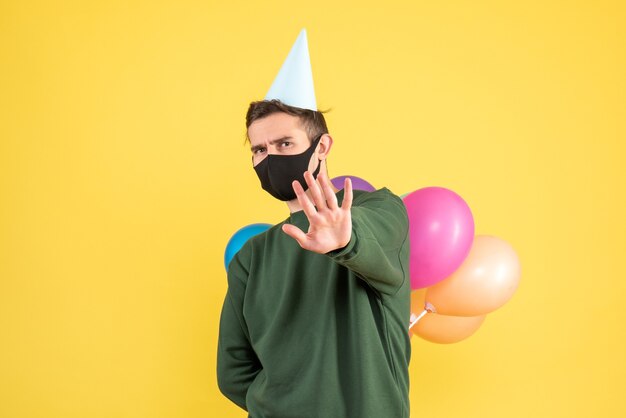 Front view young man with party cap and colorful balloons hiding balloons behind his back standing on yellow 