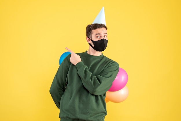 Front view young man with party cap and colorful balloons hiding his balloons behind his back standing on yellow 