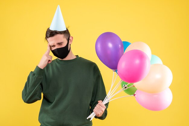 Front view young man with party cap and colorful balloons holding his head standing on yellow 