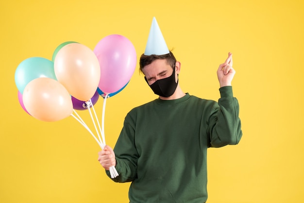 Front view young man with party cap and colorful balloons making good luck sign standing on yellow 