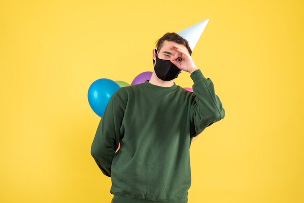 Вид спереди молодой человек с партийной кепкой и разноцветными воздушными шарами, ставя знак ОК перед его глазом, стоящим на желтом