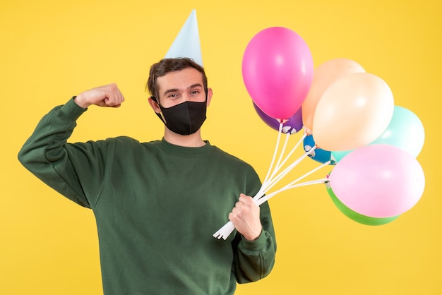 Front view young man with party cap and colorful balloons showing muscle standing on yellow 