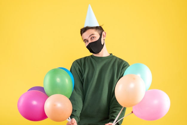 Front view young man with party cap and colorful balloons standing on yellow background copy space