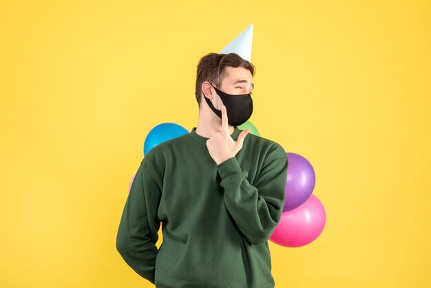 Front view young man with party cap and colorful balloons standing on yellow background free place