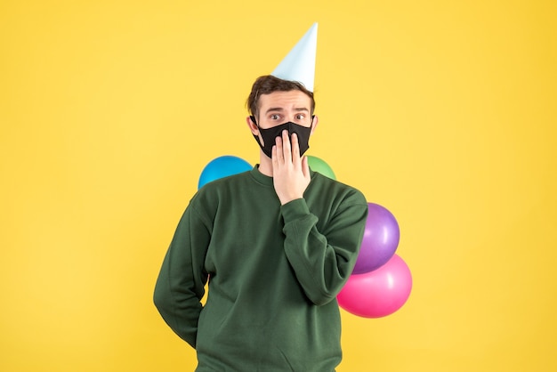 Front view young man with party cap and colorful balloons standing on yellow background