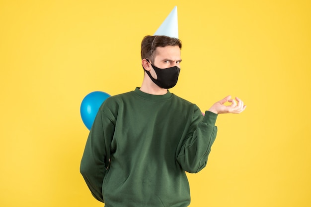 Front view young man with party cap and colorful balloons standing on yellow 