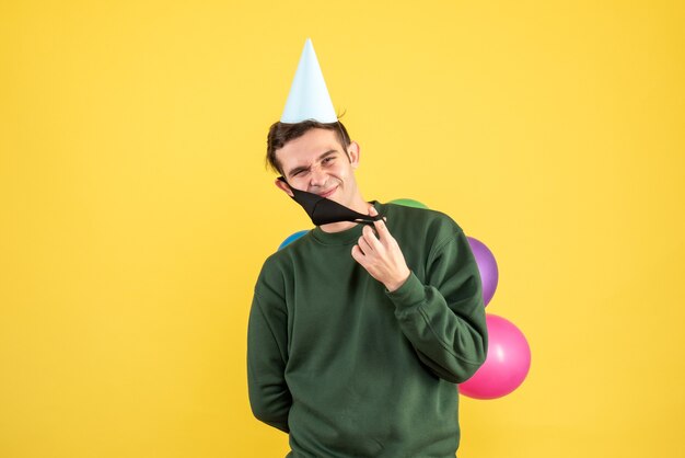 Front view young man with party cap and colorful balloons taking off his mask on yellow background