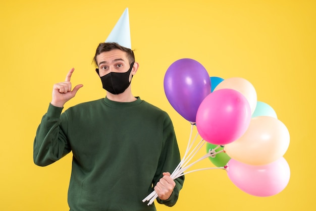 黄色の背景に黒いマスクを身に着けているパーティーキャップとカラフルな風船と正面図の若い男
