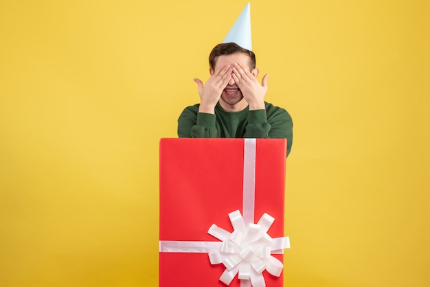 Free photo front view young man with party cap covering his eyes with hands standing behind big giftbox on yellow background