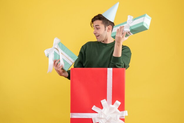 Front view young man with party cap and gifts standing behind big giftbox on yellow background