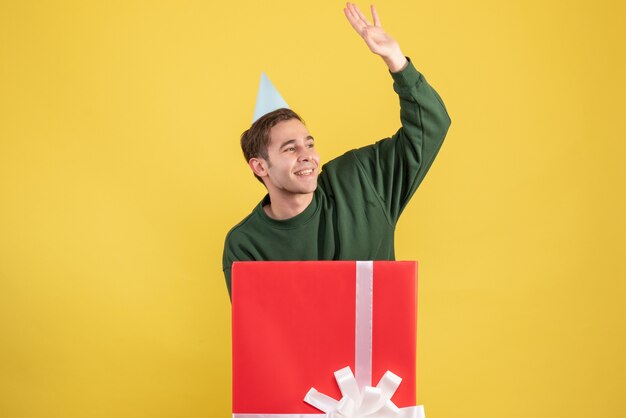 Вид спереди молодой человек в кепке приветствует кого-то, стоящего за большой подарочной коробкой на желтом фоне