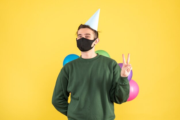 Front view young man with party cap hiding balloons behind his back standing on yellow background