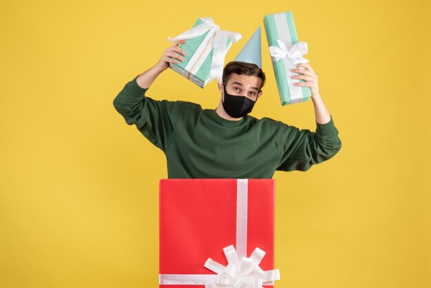 Front view young man with party cap holding gifts standing behind big giftbox on yellow background