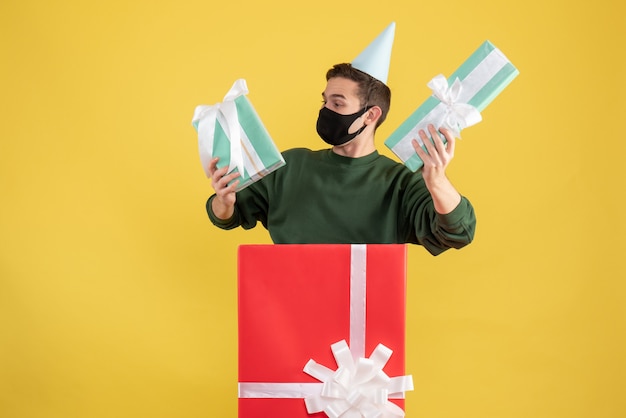 Front view young man with party cap and mask holding gifts standing behind big giftbox on yellow background