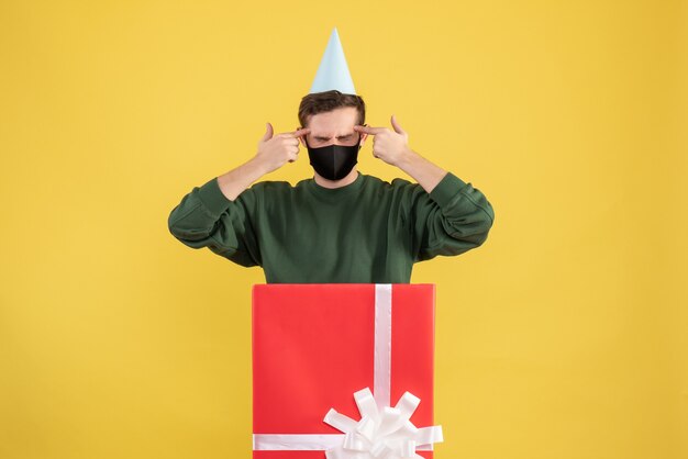 Front view young man with party cap putting fingers to his temple standing behind big giftbox on yellow background