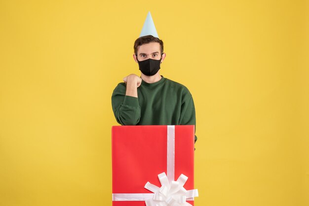 Front view young man with party cap standing behind big gift box on yellow background