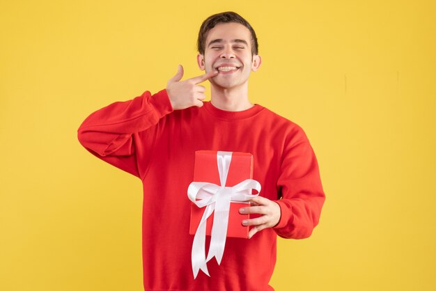 Front view young man with red gift box pointing at his smile on yellow background