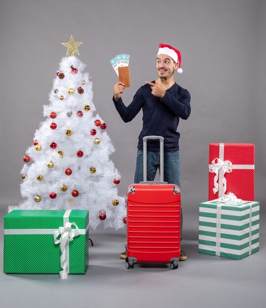 白いクリスマスツリーと灰色のプレゼントの周りに彼の旅行チケットを示す赤いスーツケースを持つ正面図の若い男