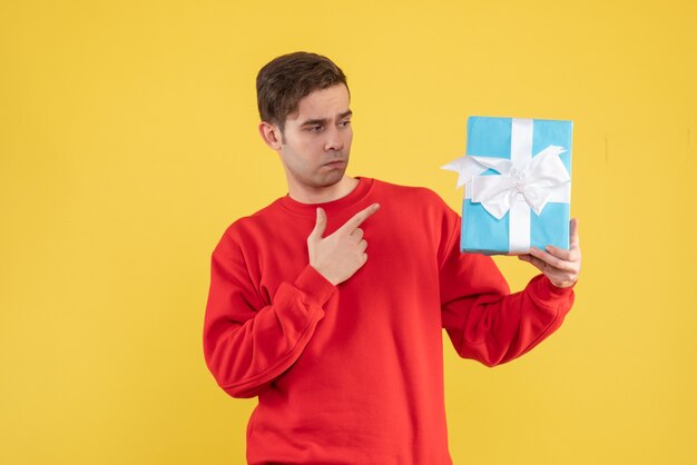 Front view young man with red sweater pointing at gift on yellow background