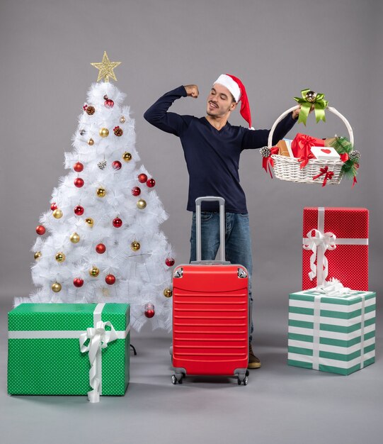 Front view young man with santa hat holding gift basket showing arm muscles near xmas tree on grey isolated