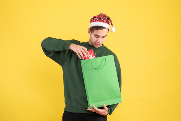 Front view young man with santa hat holding green shopping bag and gift standing on yellow background copy space