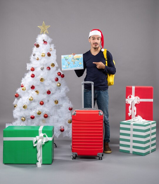 Front view young man with santa hat holding map near xmas tree on grey isolated