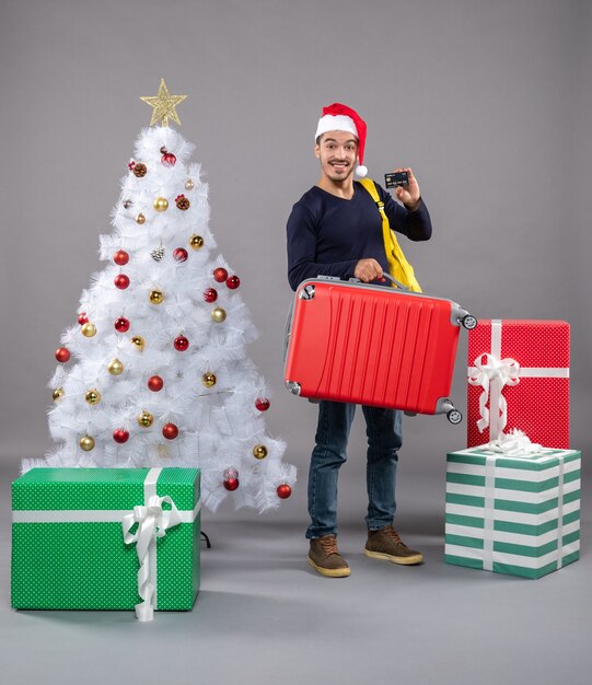 Бесплатное фото Вид спереди молодой человек в шляпе санта-клауса, держащий красный чемодан, показывает карту вокруг подарков на сером изолированном