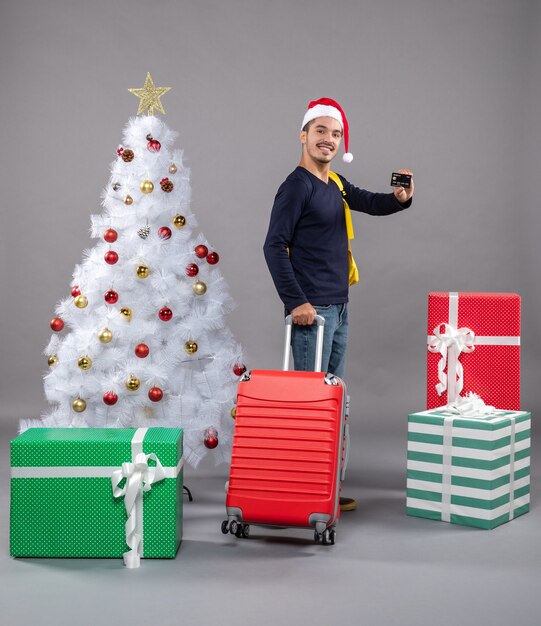 Front view young man with santa hat holding red suitcase showing a card near xmas tree on grey isolated