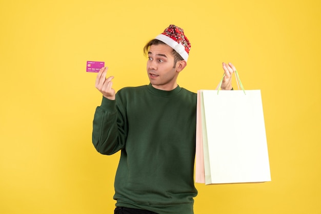 黄色の背景のコピー場所に立っている買い物袋とカードを保持しているサンタ帽子を持つ正面図の若い男