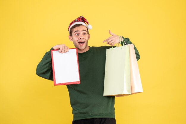 노란색 배경에 쇼핑백과 클립 보드를 들고 산타 모자와 함께 전면보기 젊은 남자