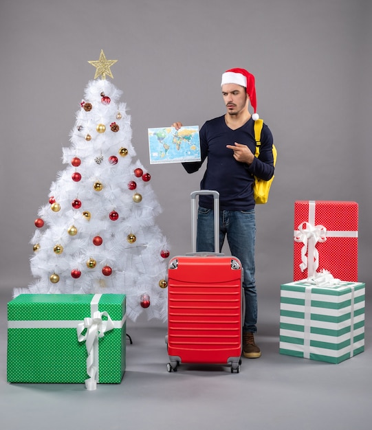 Front view young man with santa hat holding world map standing near xmas tree on grey isolated