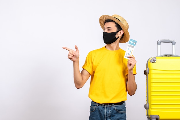 Front view young man with straw hat and mask standing near yellow suitcase holding travel ticket pointing at left