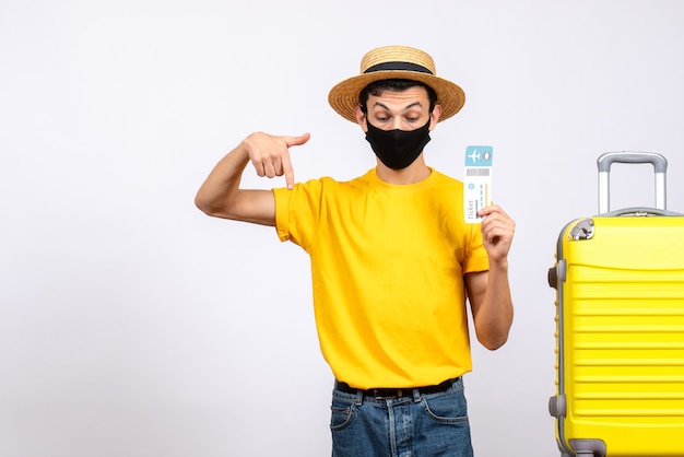 Front view young man with straw hat and mask standing near yellow suitcase holding travel ticket pointing at below