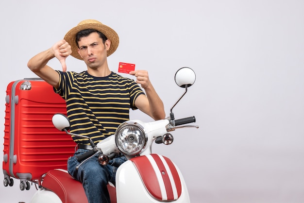 front view of young man with straw hat on moped holding credit card giving thumb down