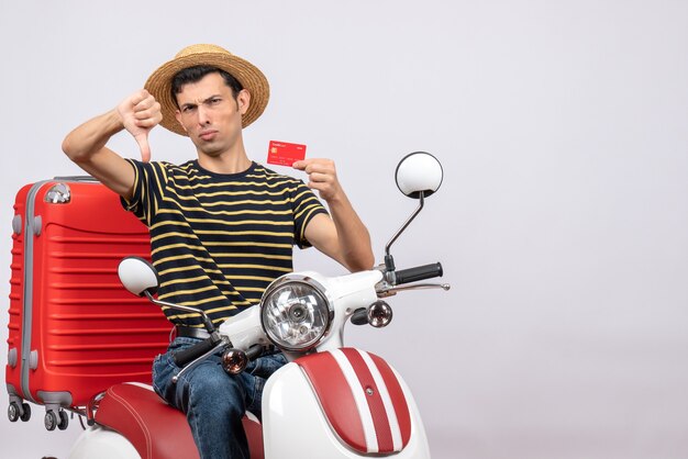 Front view of young man with straw hat on moped holding credit card giving thumb down