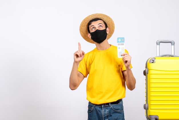 Вид спереди молодой человек в соломенной шляпе, стоящий возле желтого чемодана с билетом на самолет
