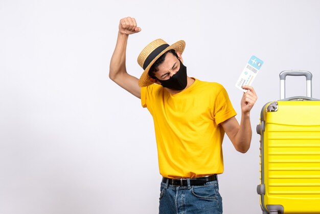 Front view young man with straw hat standing near yellow suitcase holding travel ticket expressing his feelings