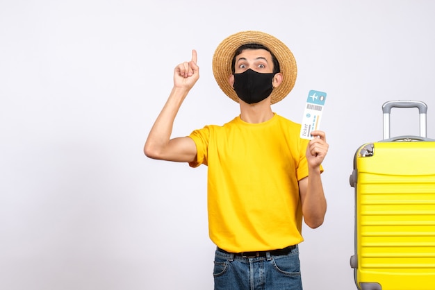 Front view young man with straw hat standing near yellow suitcase holding travel ticket pointing finger up