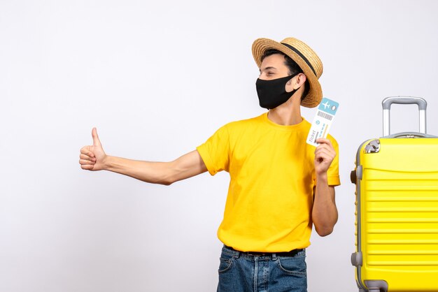 가입 엄지 손가락을 가리키는 여행 티켓을 들고 노란색 가방 근처에 서 밀 짚 모자와 전면보기 젊은 남자