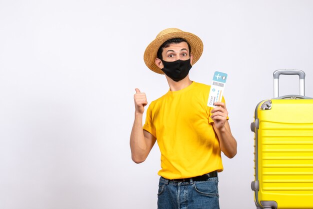 Front view young man with straw hat standing near yellow suitcase holding travel ticket pointing at behind