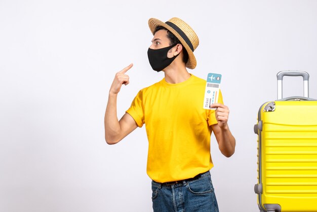 Front view young man with straw hat and yellow t-shirt standing near yellow suitcase holding plane ticket