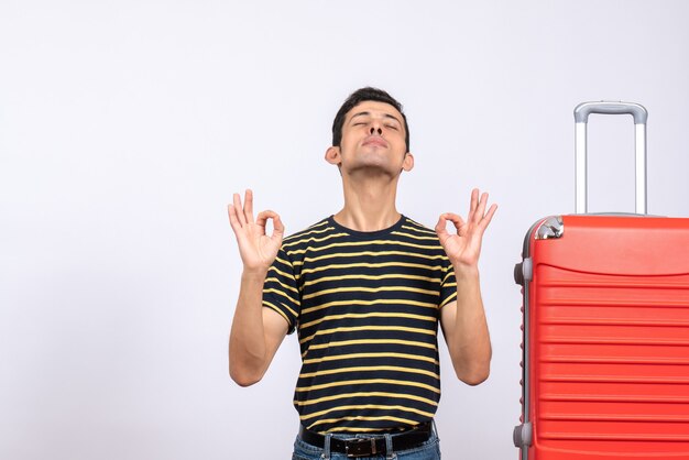Вид спереди молодой человек с полосатой футболкой и красным чемоданом делает знак ОК, закрывая глаза