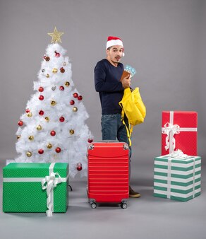 旅行​チケット​と​赤い​スーツケース​を​持った​正面図​の​若い​男​が​灰色​の​孤立した​さまざまな​プレゼント​の​周り​に​彼​の​黄色​の​バック​パック​を​チェック