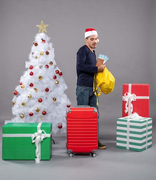 Front view young man with travel ticket and red suitcase checking his backpack around presents on grey