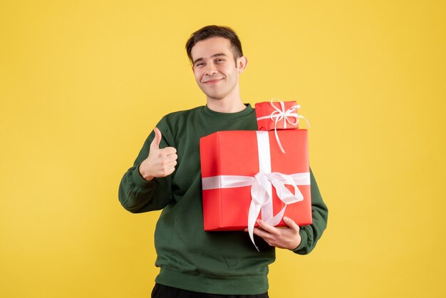 Front view young man with xmas gift making thumb up sign standing on yellow background