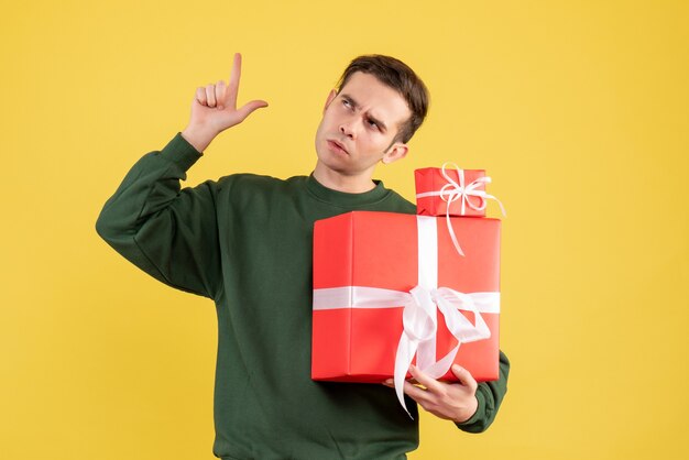 Front view young man with xmas gift pointing at ceiling standing on yellow background