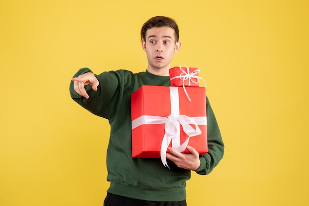 Front view young man with xmas gift pointing at something standing on yellow background